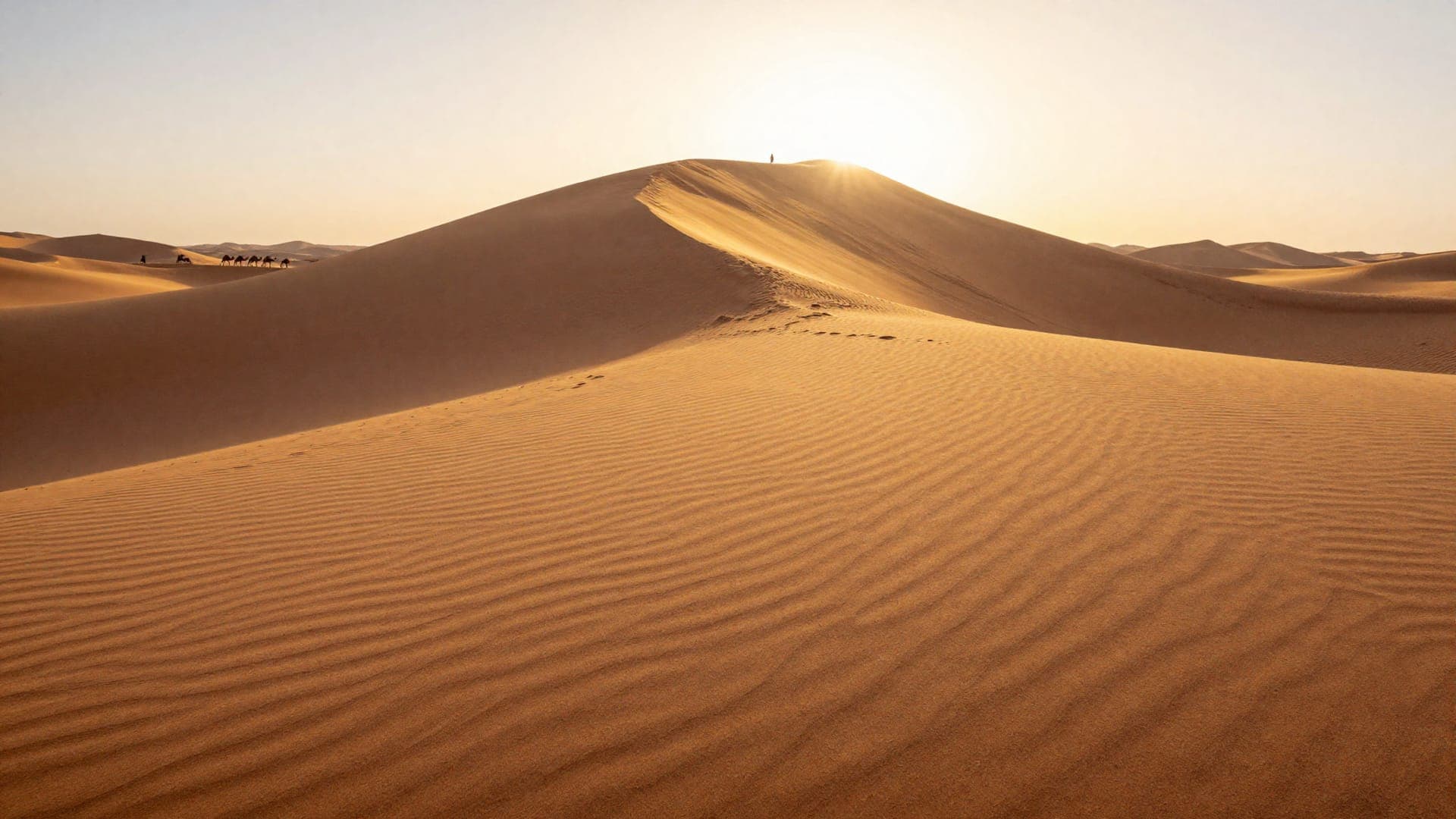 The sun breaking over a high dune ridge, a single figure standing on the crest against it, and a camel train crossing the sand far to the left.