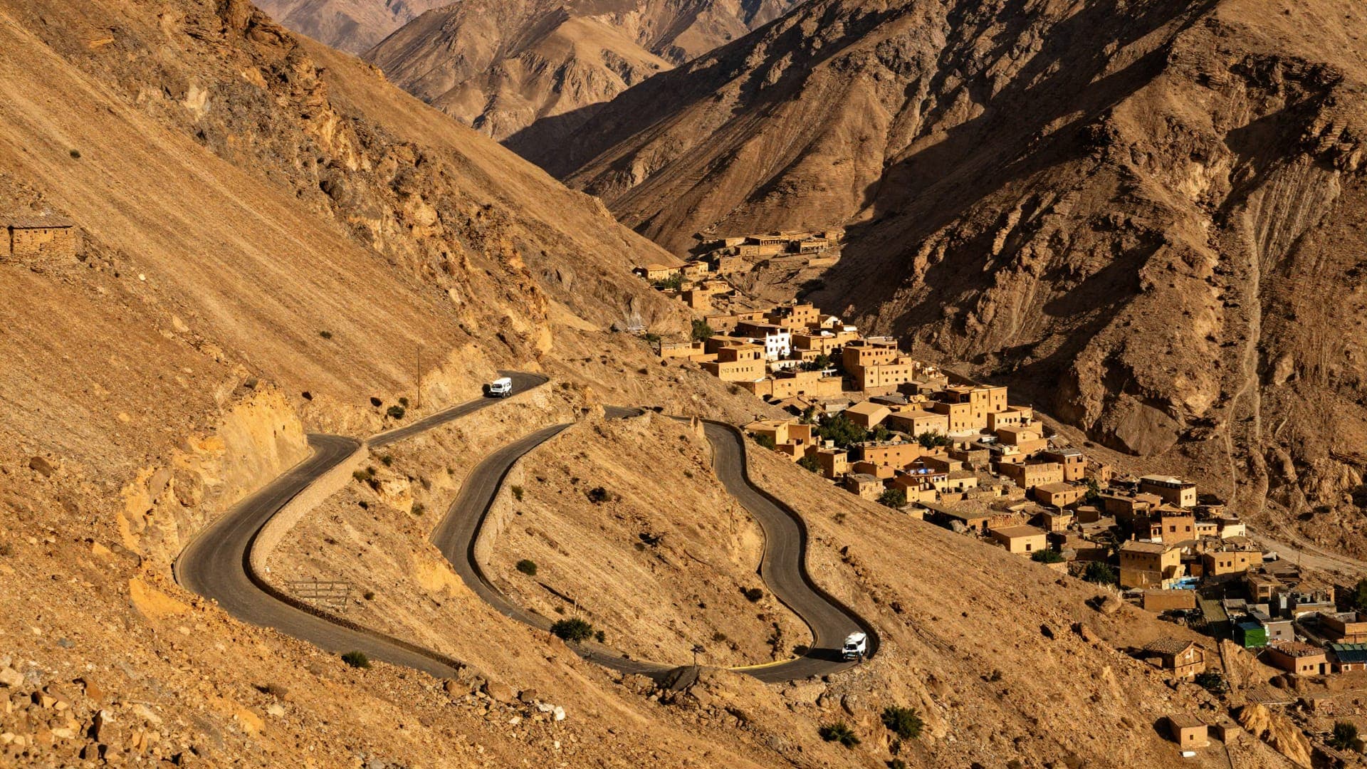 A tarmac road folding through three hairpins down a steep ochre mountainside, two white minibuses on the bends, and a village of earth-built houses terraced into the valley below.